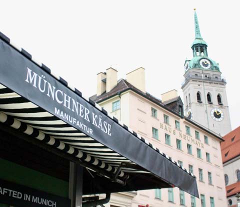 Der Stand der Münchner Käse Manufaktur am Viktualienmarkt. Im Hintergrund die Kirch St. Peter.