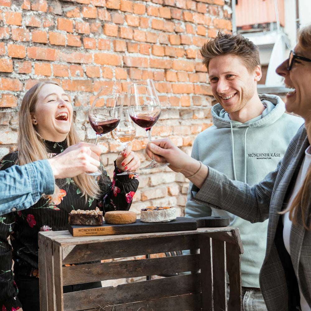 Zwei Frauen und ein Mann stoßen mit Wein an während unter den Weingläsern ein Brett mit Käse steht.