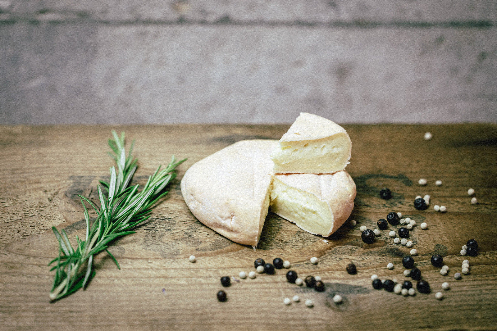 Der Münchner Isarbazi von der Münchner Käse Manufaktur liegt neben einigen Pfefferkörnern auf einer Unterlage aus Holz. Links daneben ein Zweig Rosmarin.