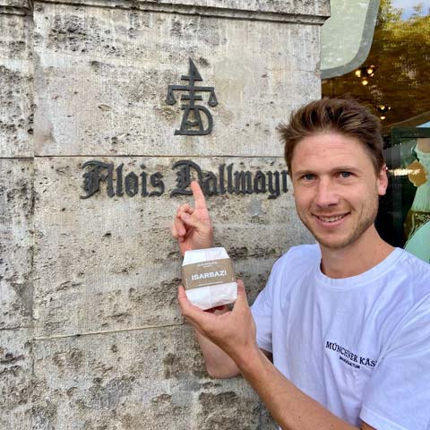 Sebastian steht mit einem verpackten Käse vor dem Logo von Alois Dallmayr