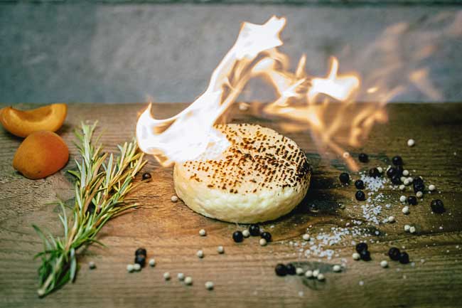 Zu sehen ist Grillkäse der Münchner Käse Manufaktur. Er liegt auf einem Holzbrett. Es sind Flammen zum Rösten zu sehen.