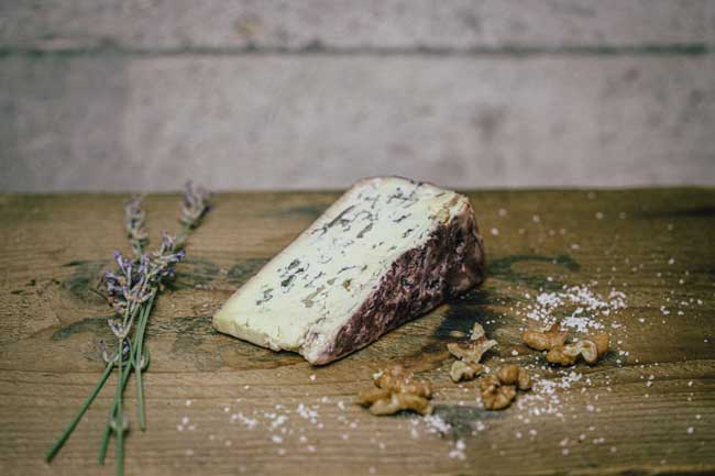 Zu sehen ist der Blaue Portugiese der Münchner Käse Manufaktur. Er liegt auf einem Holzbrett und ist angeschnitten. Es ist ein Blauschimmelkäse mit Portwein affiniert.