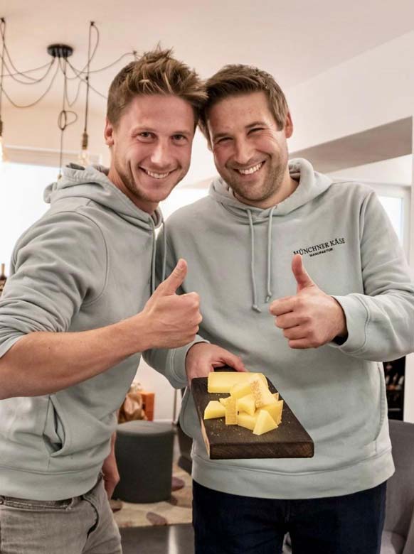 Zwei gründer der Münchner Käse Manufaktur heben den Daumen hoch und haben ein Brett mit Käse vor sich. Sie lachen.