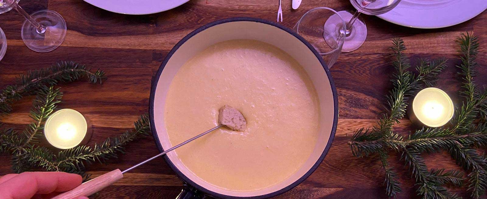 Ein Stück Brot wir aufgespießt auf einer Gabel in einen gusseisernen Topf mit Fondue getunkt.
