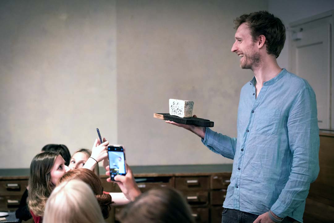 Johannes praesentiert Kaese vor einer Gruppe waehrend eine Teilnehmerin den Kaese mit einem Handy fotorafiert