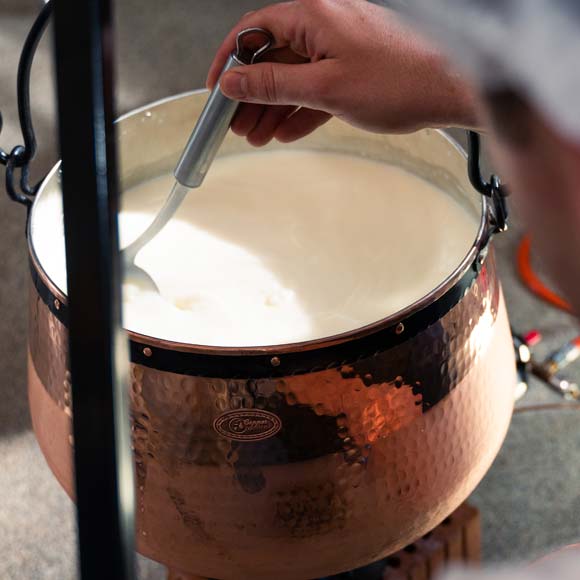Ein Kupferkessel mit Milch zum Käse machen wird mit einer Schöpfkelle umgerührt.