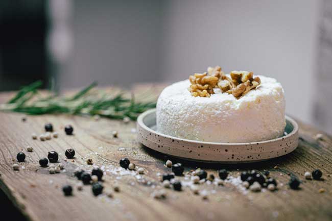 Zu sehen ist Münchner Ricotta der Käse der Münchner Käse Manufaktur. Er liegt in einem kleinen Schüsselchen auf einem Holzbrett. Als Deko werden Walnüsse verwendet.